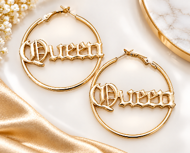 A pair of gold hoop earrings with the word 'Queen' in a gothic font, styled on a white marble surface with gold fabric and small white flowers.
